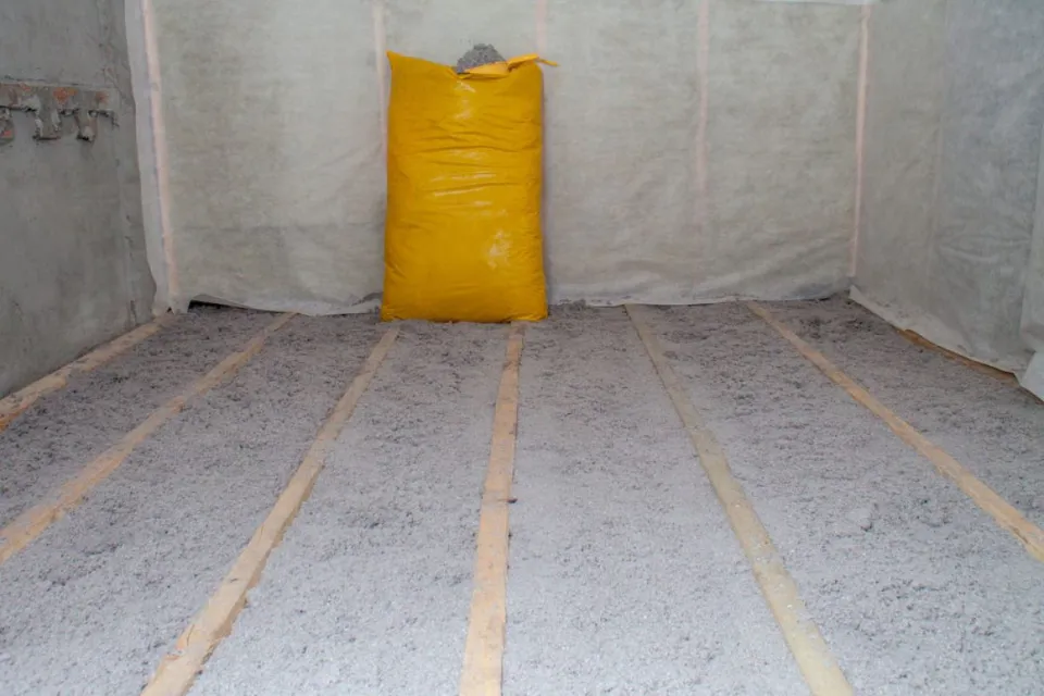  Loose-fill cellulose insulation installed between floor joists, with a yellow bag of insulation material in a residential construction space.